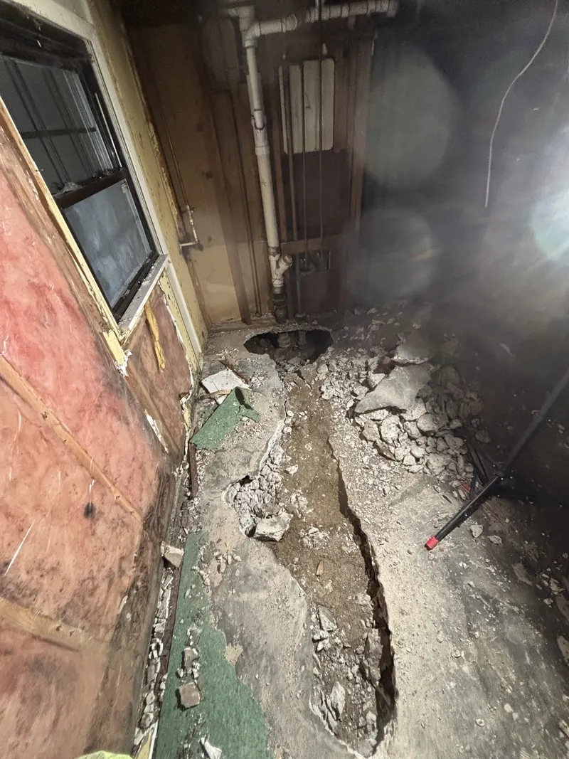 Narrow excavation beside a foundation exposing below-grade piping and soil conditions.